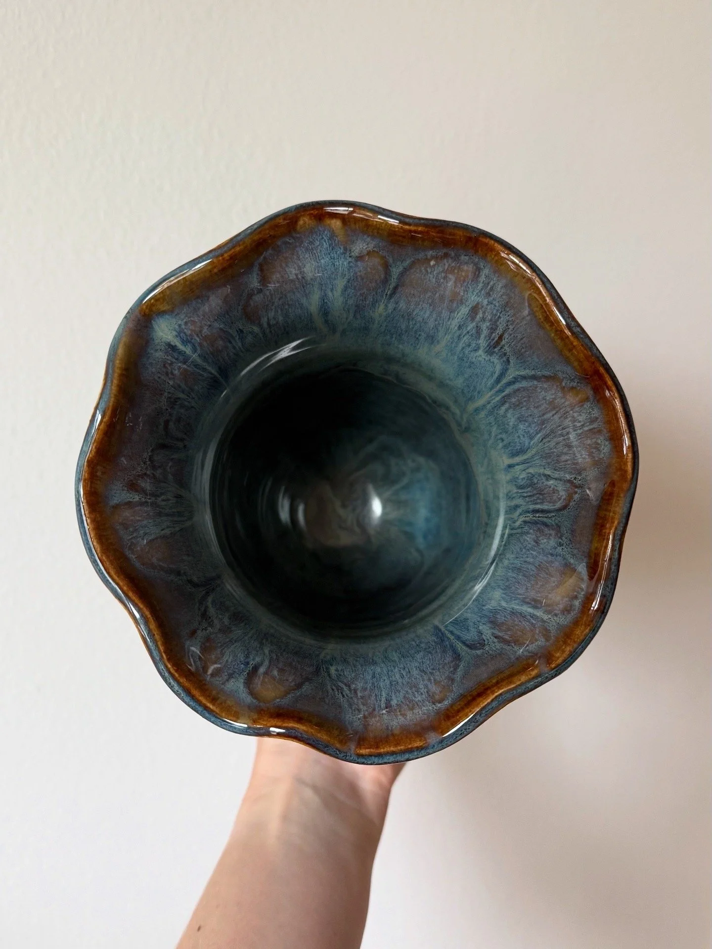 Top view of an artisan ceramic vase with a wavy rim, glazed in blue and brown tones, held in a person's hand against a plain white background.