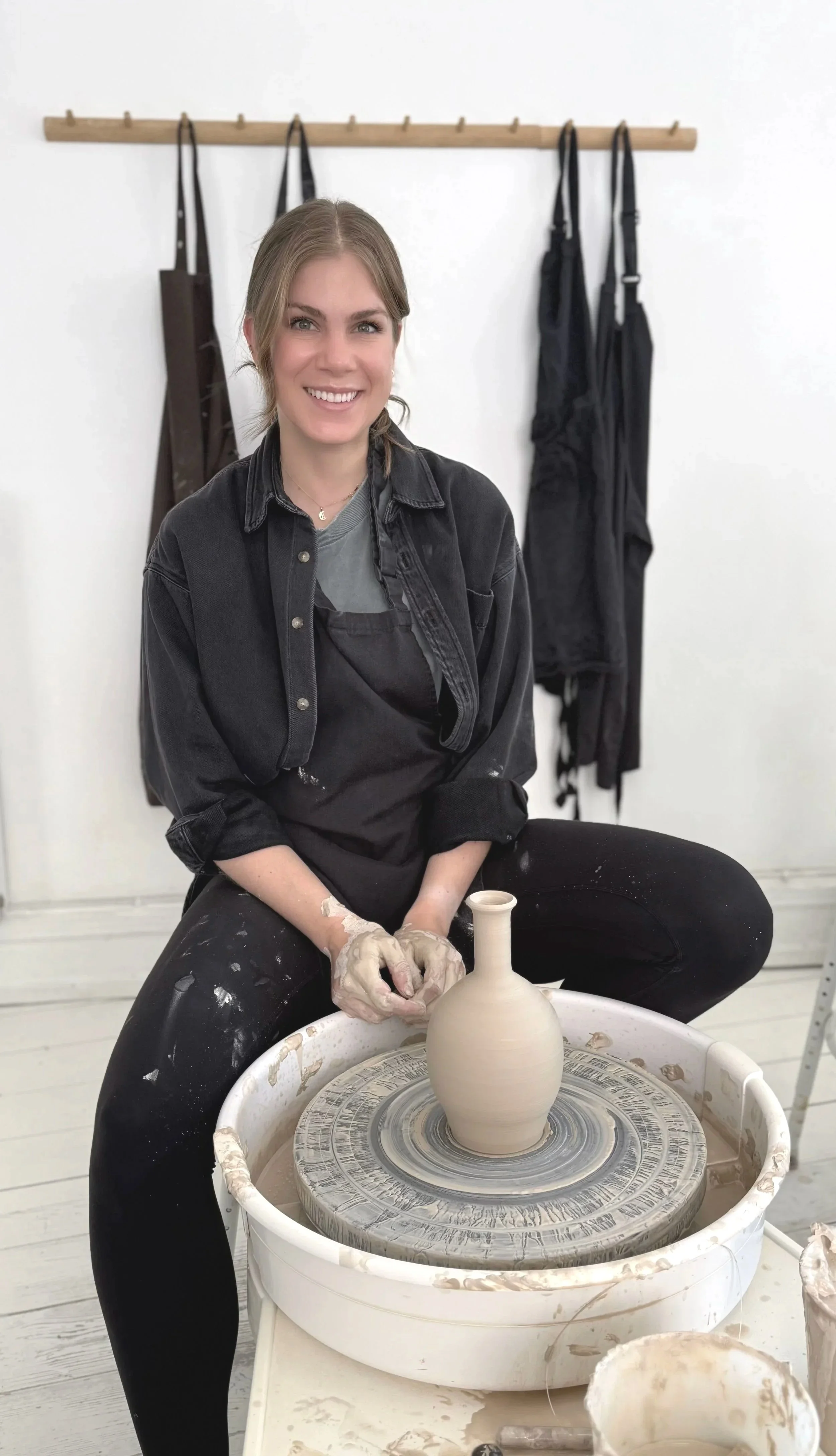 Young woman smiling while working on pottery at a wheel, with ceramic vase in progress. She is sitting in a pottery studio.