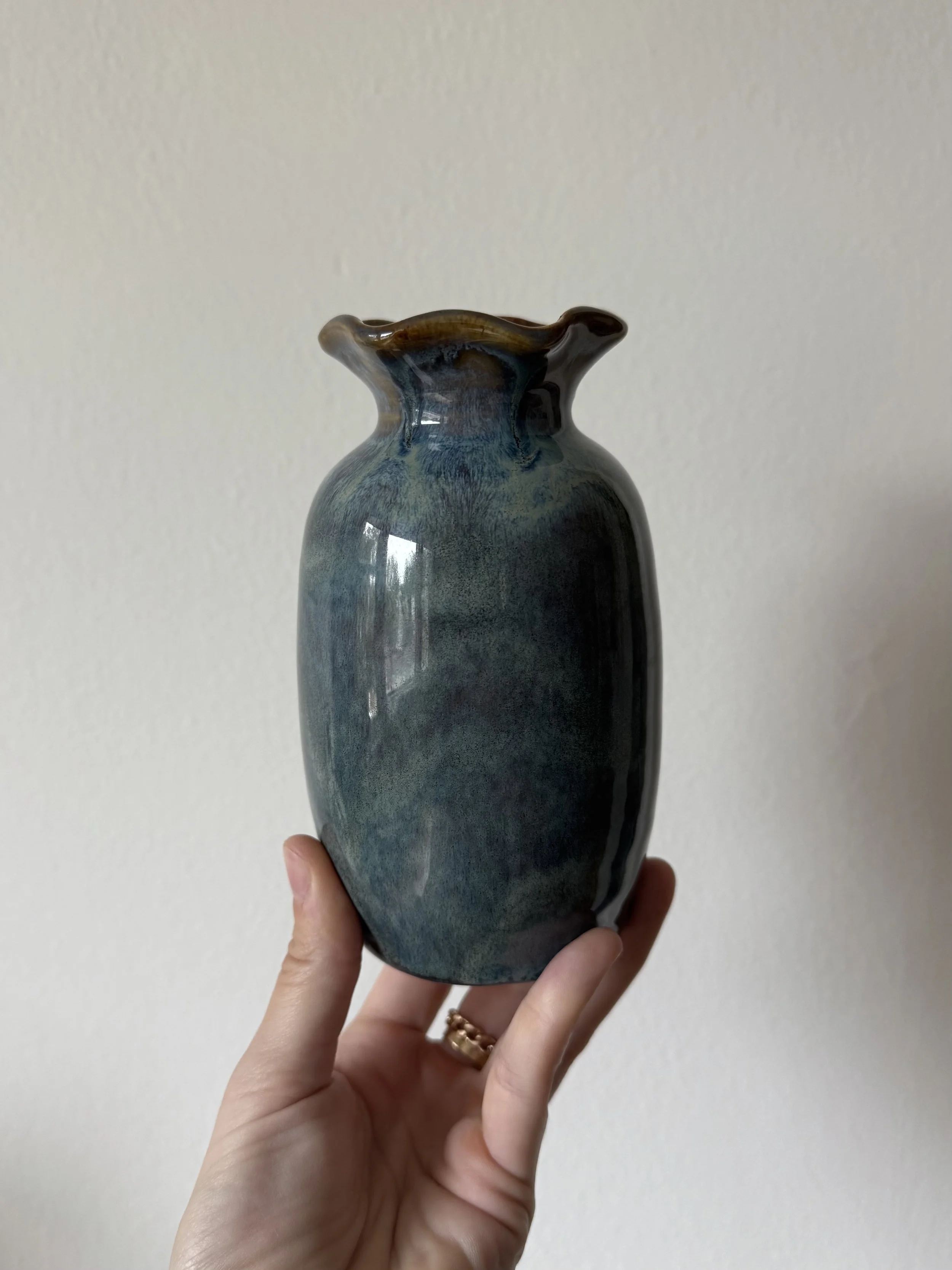 A person holding a ceramic vase with a narrow neck, wide body, and ruffled top edge, glazed in shades of blue, and brown.