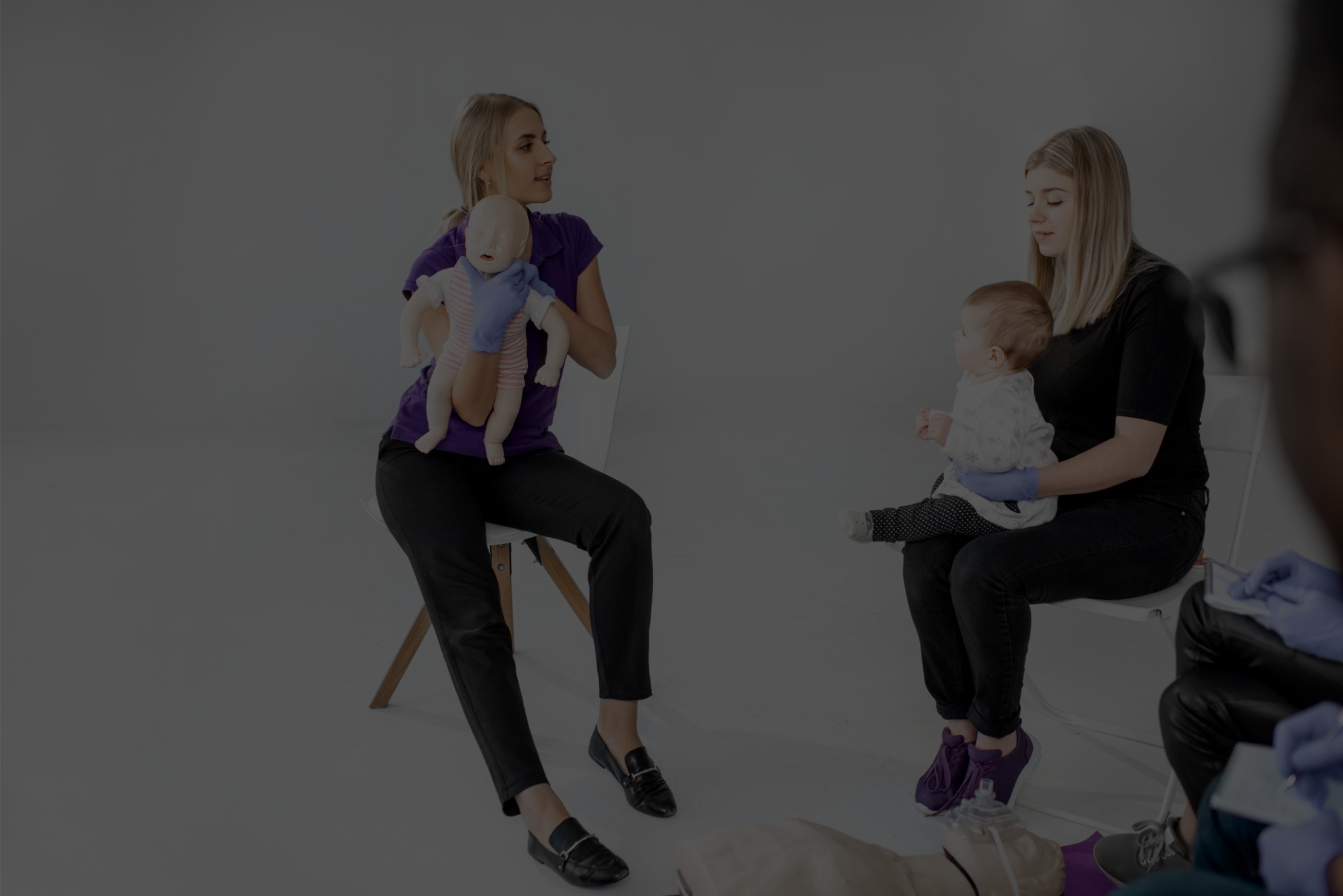 Two women, one holding a lifelike baby doll and the other holding a real baby, in a medical setting with healthcare professionals, practicing infant care techniques.