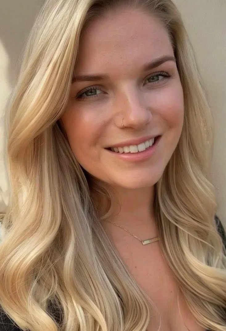 Close-up photo of a young woman with long, blonde, wavy hair, fair skin, light-colored eyes, and a nose piercing, smiling at the camera.
