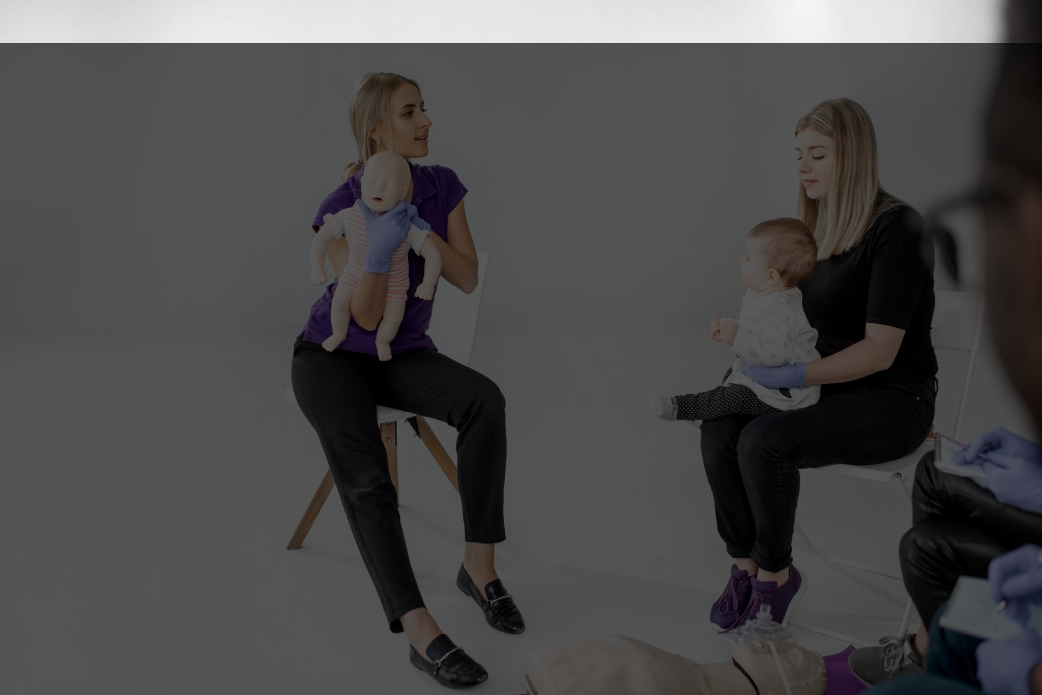 Two women, one in a purple shirt and black pants holding a baby doll, and the other in a black shirt and jeans sitting on a chair with a young girl, are in a medical setting. The woman in black appears to be a healthcare professional, wearing gloves, and the woman in purple is a patient. The healthcare professional is preparing medical supplies.