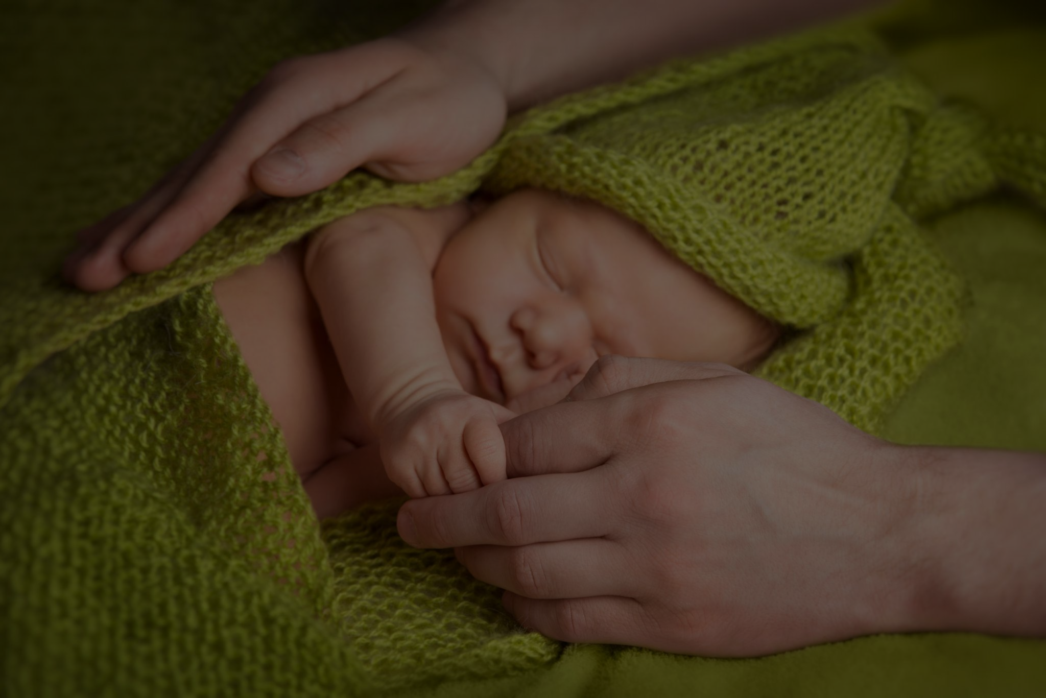 Close-up of an adult hand gently holding a tiny baby's hand, wrapped in a green knitted blanket.