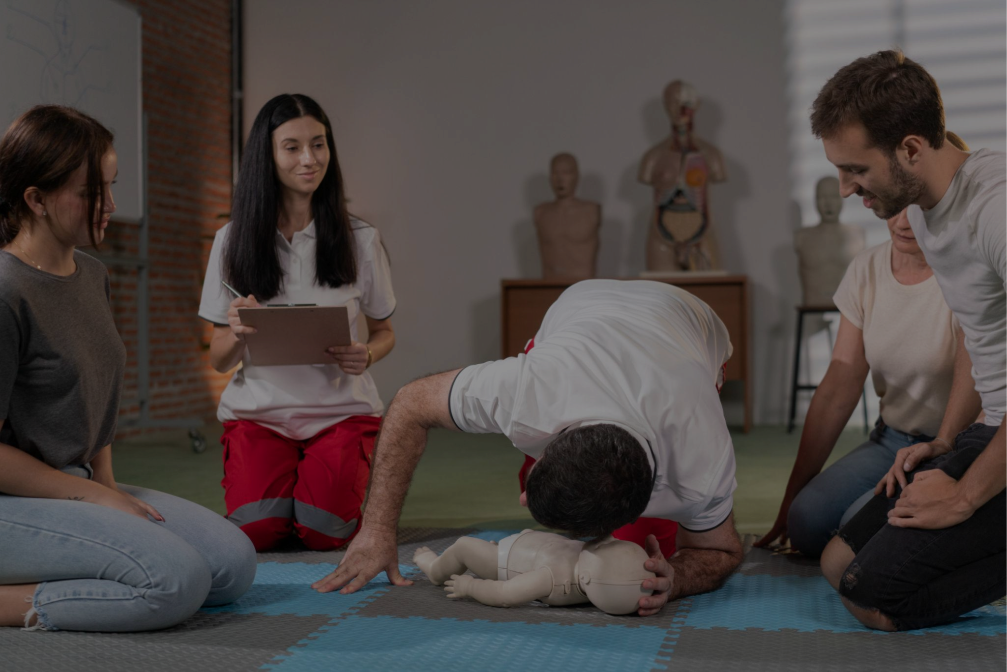A group of people participating in a CPR training class indoors, practicing on a CPR mannequin with medical anatomical models in the background.
