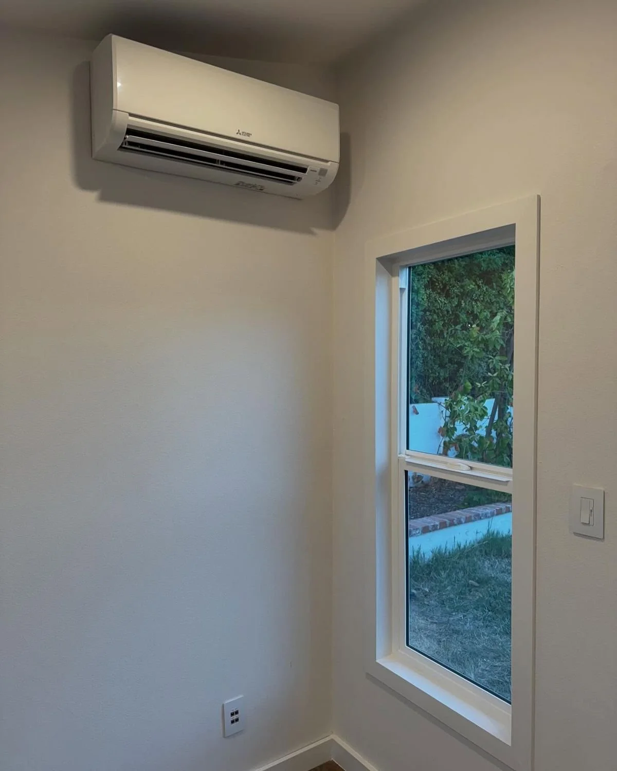 A room corner with a white wall-mounted air conditioning unit, a window with a view of green bushes outside, and a white switch on the wall.