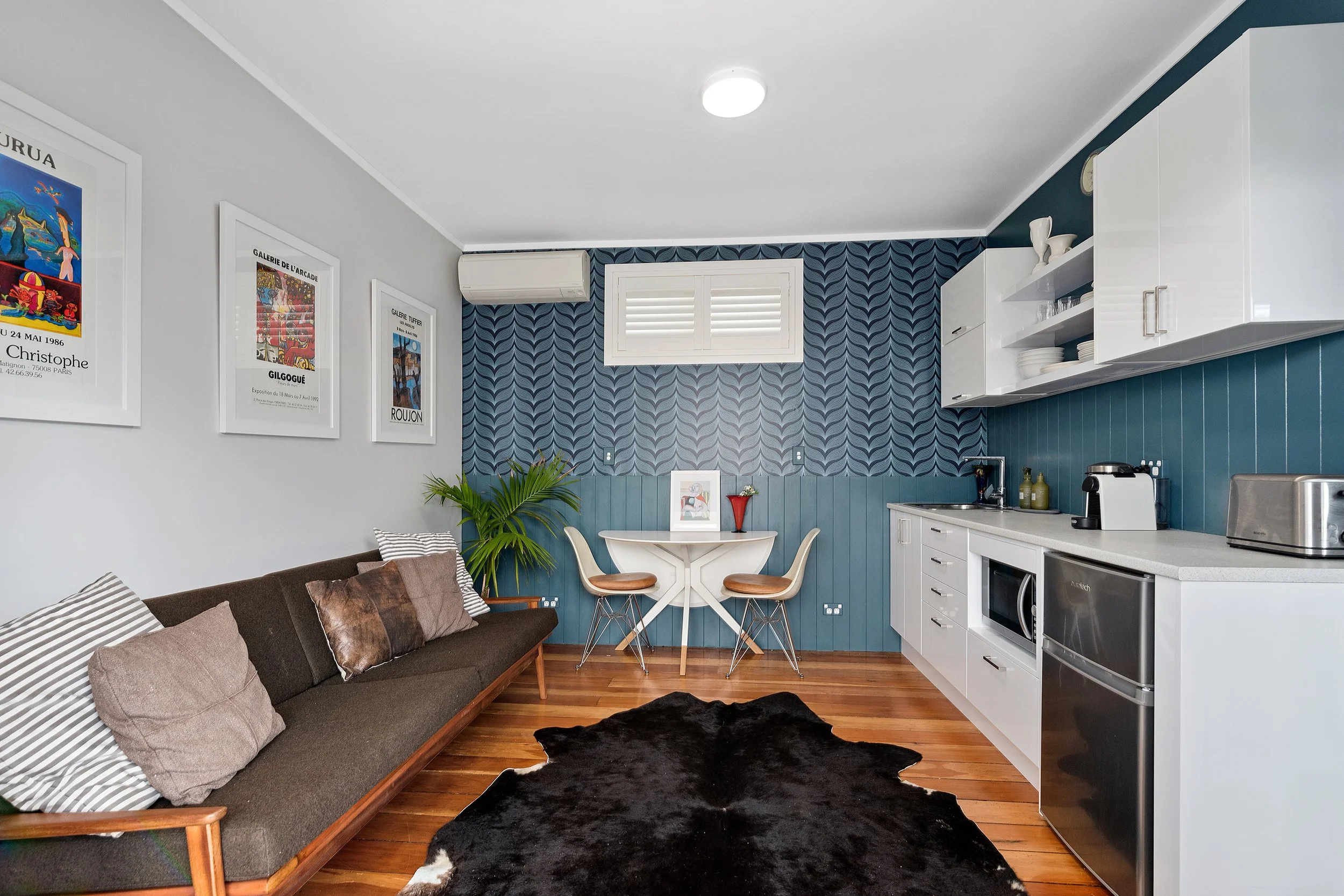 Living room with a brown sofa, decorative pillows, a black and white cowhide rug, a blue accent wall with wall art, white kitchen cabinets, a small window with shutters, and wall-mounted heat pump installed destreetly in a modern living room.