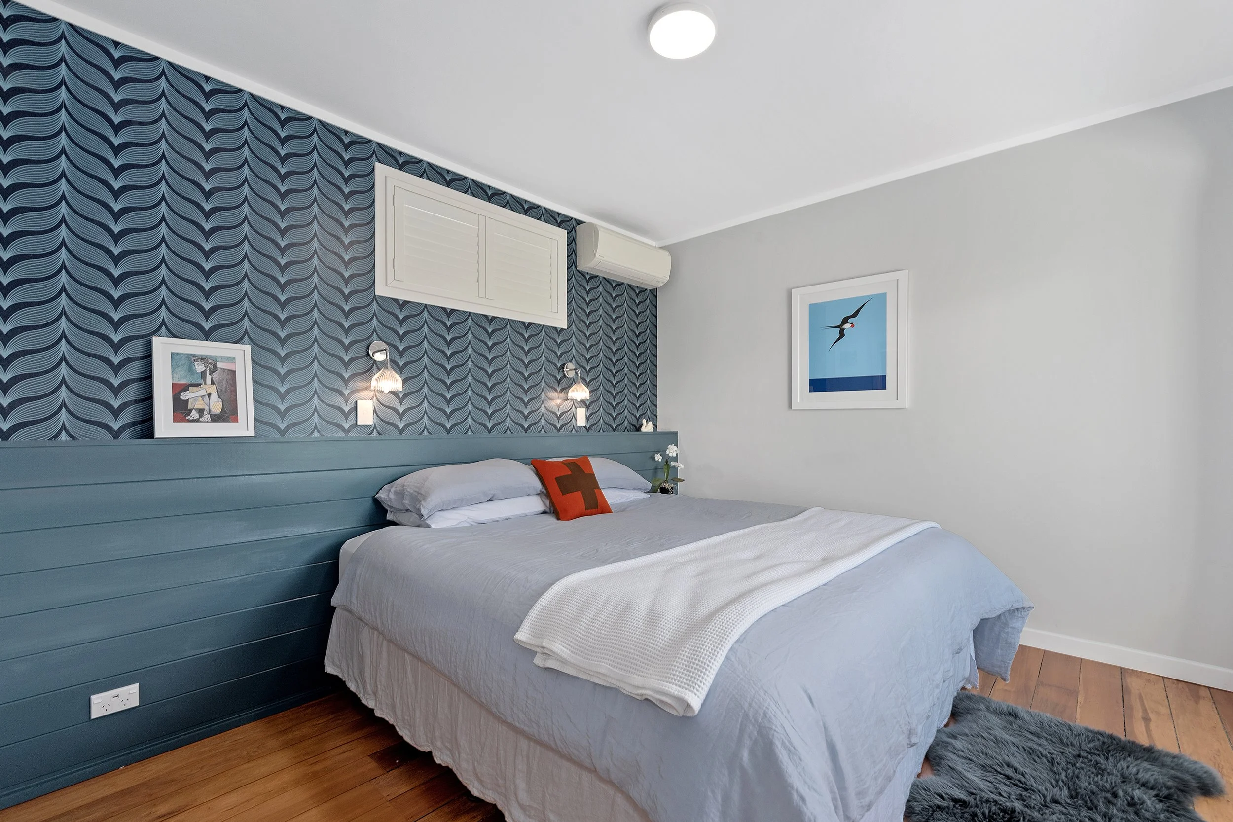 A neatly made bed with gray bedding and a decorative orange pillow, set against a blue accent wall with a modern geometric pattern, artwork on the wall, and a furry gray rug on a wooden floor. White wall-mounted heat pump installed discreetly.