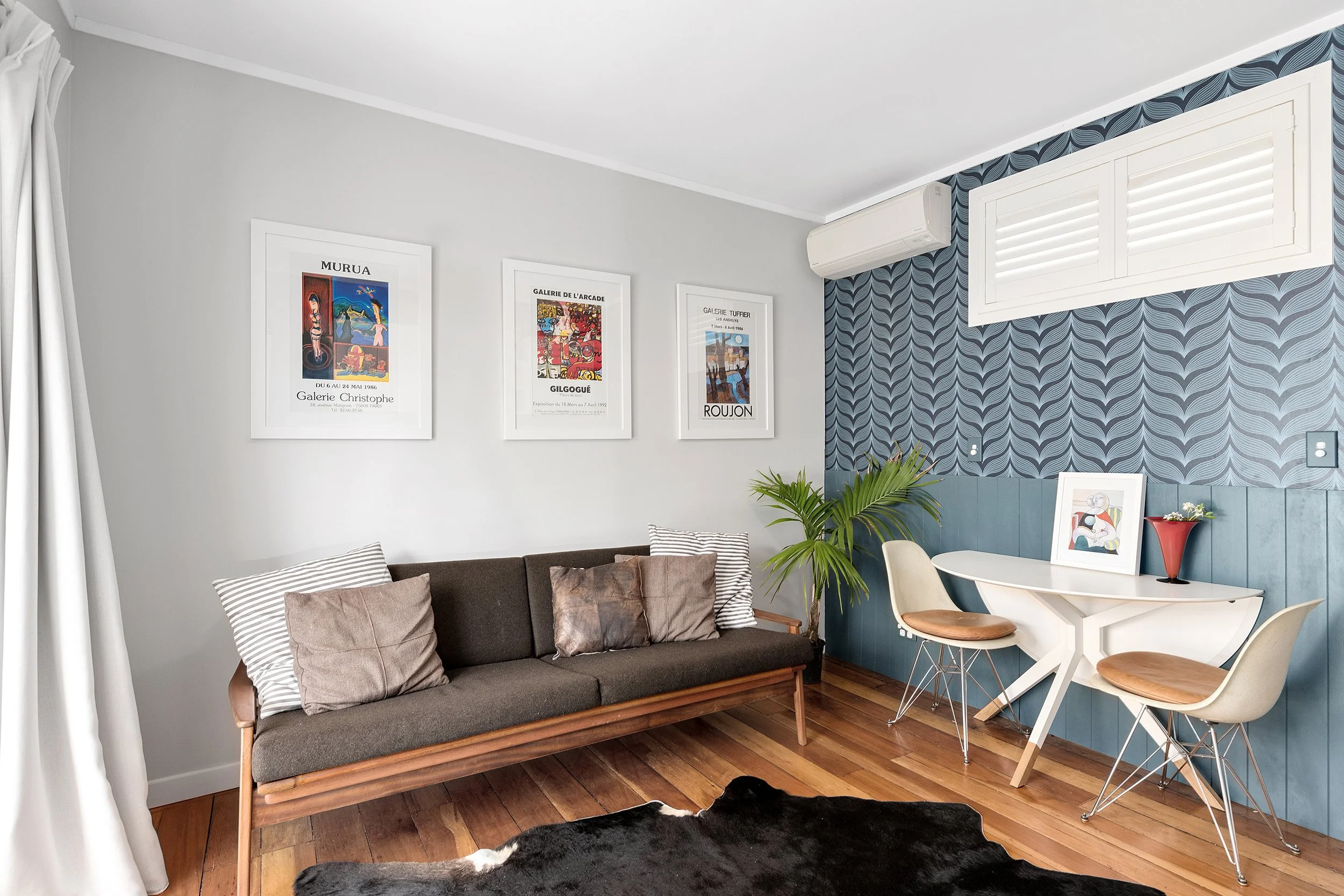 A living room with a brown sofa, pillows, wall art, a piano, a table with chairs, plants, and a window with a shutter, decorated in a modern style white wall-mounted heat pump installed discreetly in corner of room.
