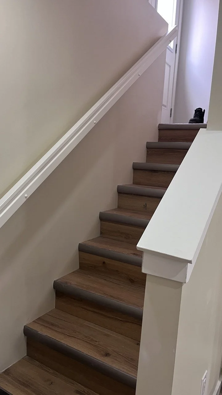 Wooden staircase with a white handrail and beige walls, leading up to a door at the top, with a black shoe or boot on the floor near the door.