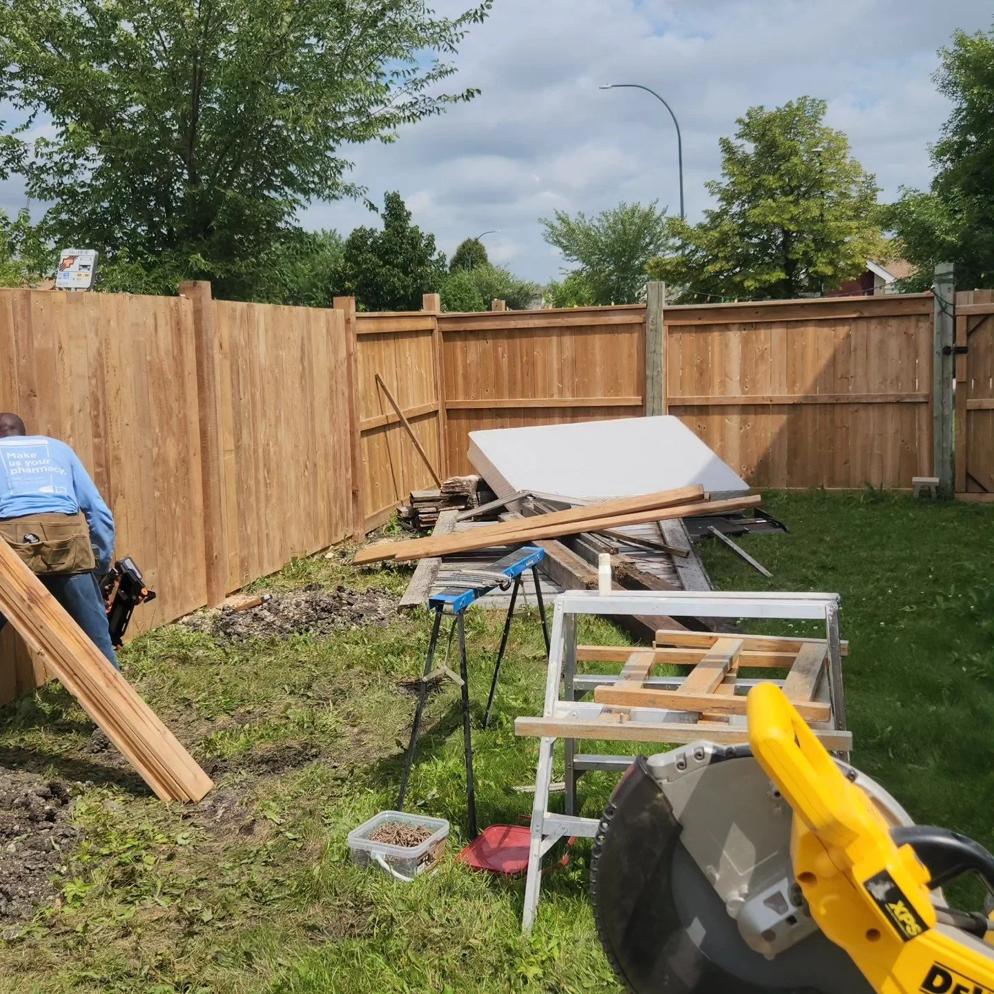 Backyard fencing installation with wooden panels, construction tools, and materials scattered, including a miter saw in the foreground and a person working on the left.
