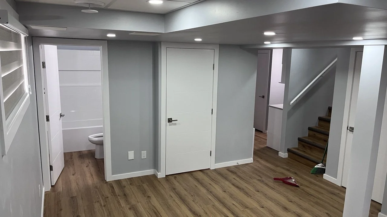 Interior basement room with wood floors, gray walls, and recessed lighting. An open door reveals a small bathroom with a toilet. A staircase with wooden steps and a white handrail leads upstairs. A broom and dustpan are on the floor.