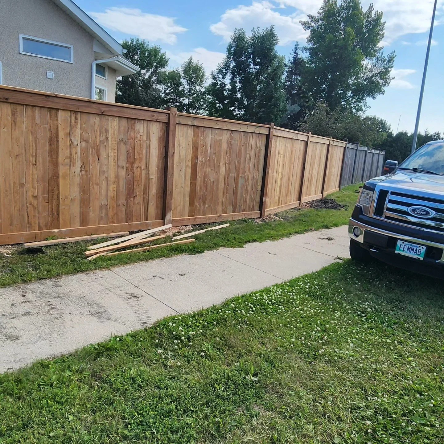 A wooden fence under construction along a sidewalk, with a black Ford truck parked nearby. The background shows a house, trees, and a blue sky with clouds.