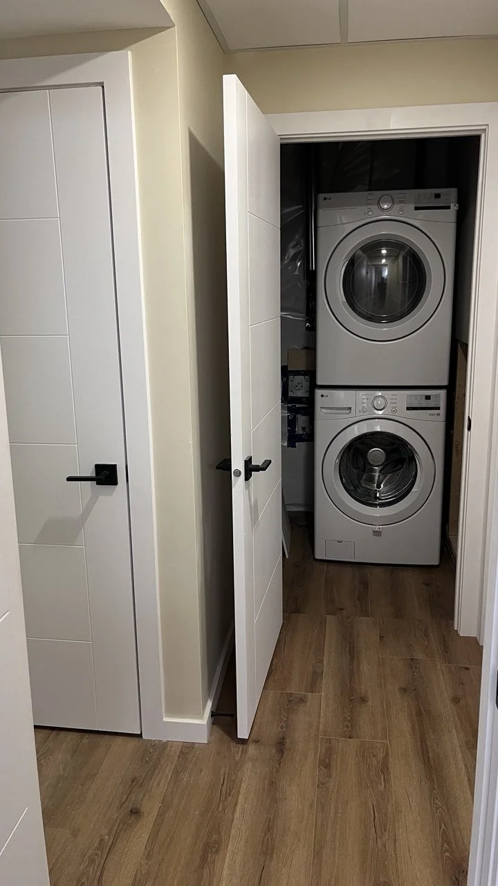 A small laundry closet with a stacked washing machine and dryer behind a partially open door, located in a room with wood flooring and white walls.