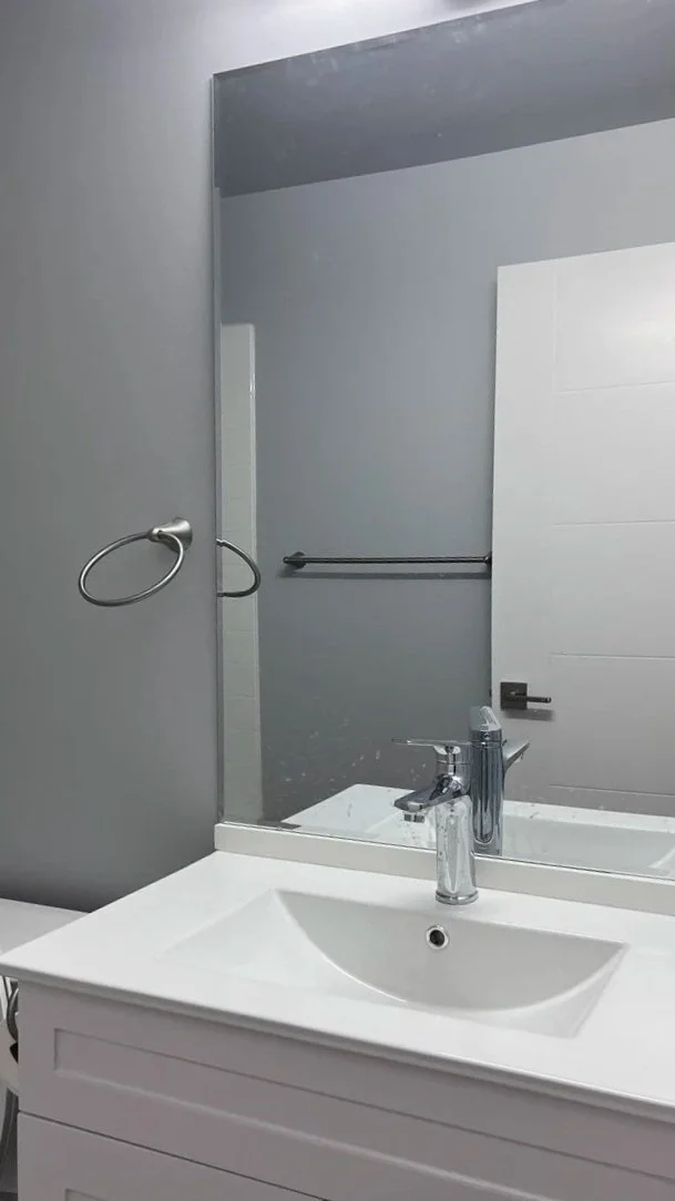 A bathroom sink with a mirror above, a towel ring to the left, and a towel bar reflected in the mirror. The bathroom has gray walls and a white door with a black door handle.