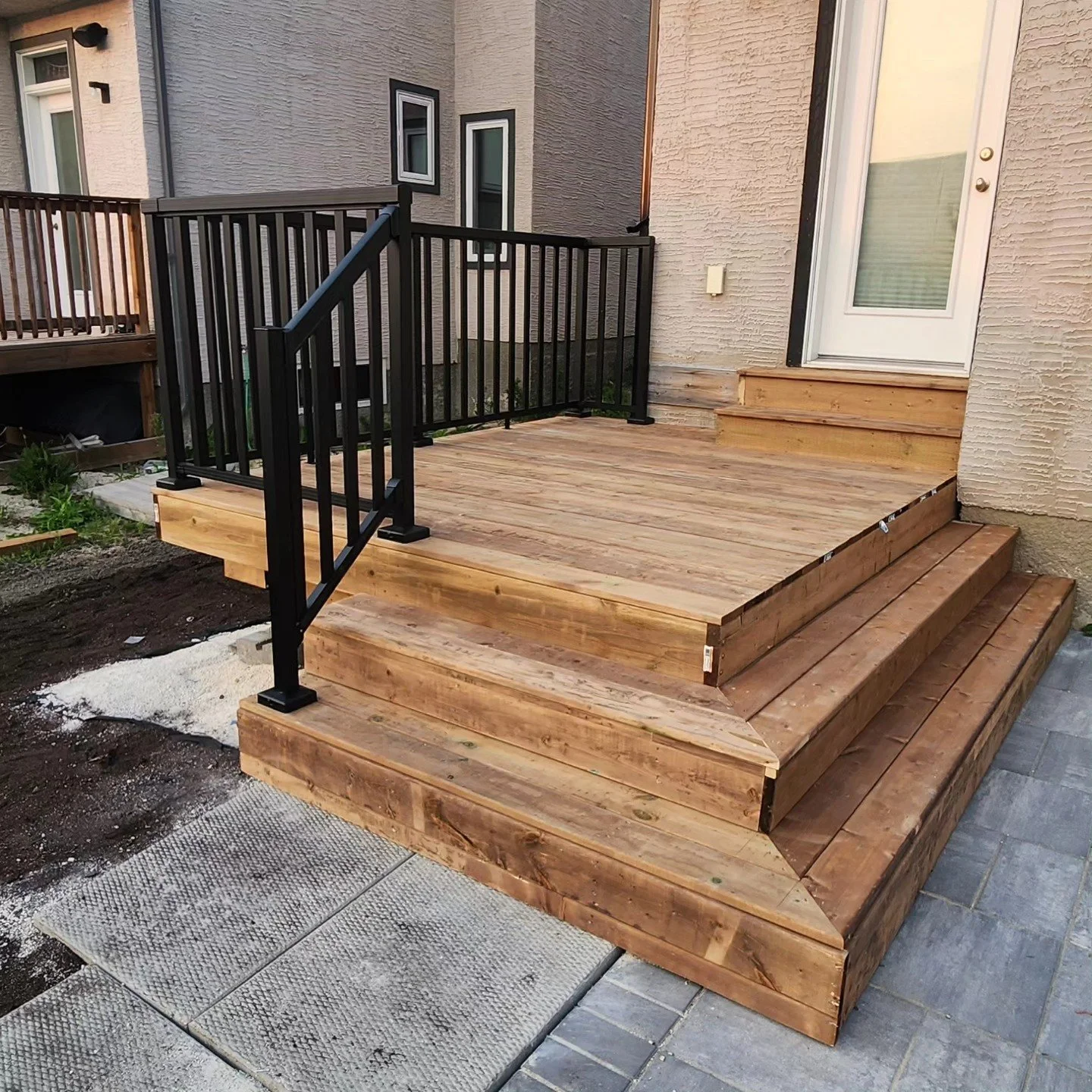 Newly built wooden deck with steps and black metal railing attached to a house with a white door and beige textured wall.
