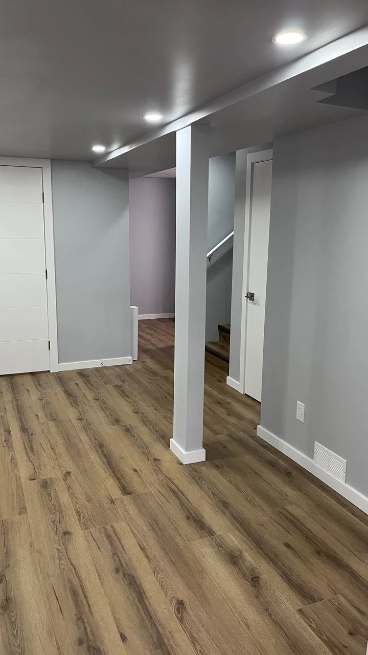 Interior view of a basement room with wooden floors, gray painted walls, white trim, recessed ceiling lights, a staircase, and a door.