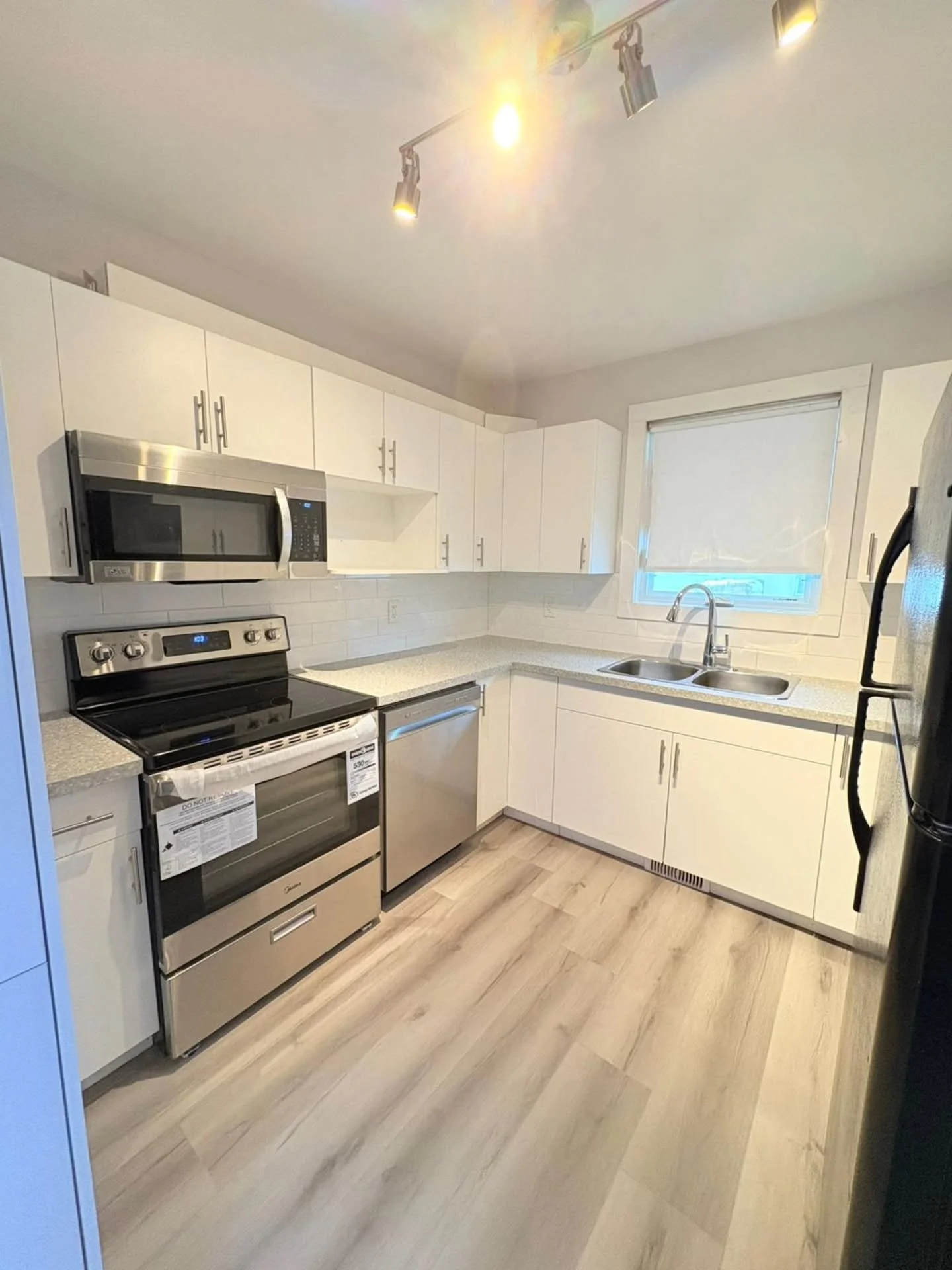 Modern kitchen with white cabinets, stainless steel appliances, a window with a shade, and wood-look flooring.