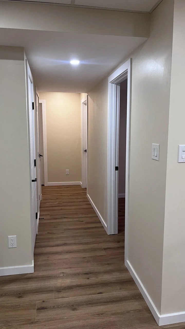 Empty hallway in a house with beige walls, hardwood flooring, and white door frames, leading to several closed doors.