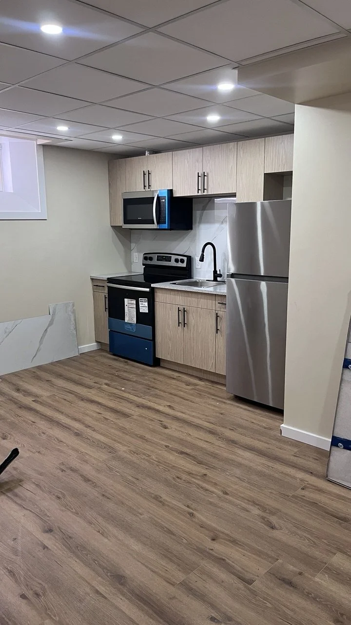 A small kitchen with light wood cabinets, a stainless steel refrigerator, black stove, black microwave, and a white sink with a black faucet. The floor is wood, and there is a small window on the left.
