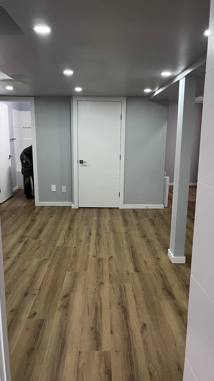 Empty room with wood laminate floors, gray walls, white door at the center, and recessed ceiling lights. Part of a man is visible on the left side near an open door.