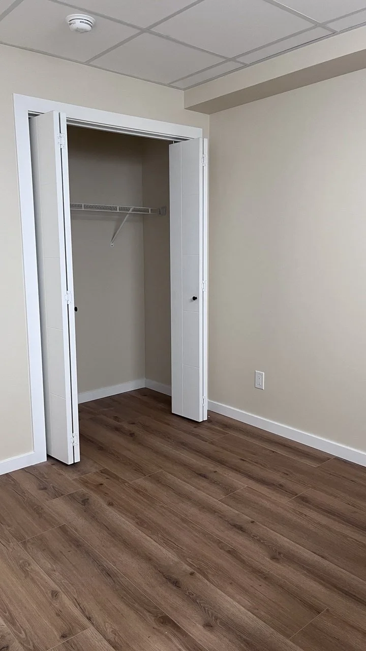Empty closet with white bi-fold doors in a room with beige walls and wooden floor.