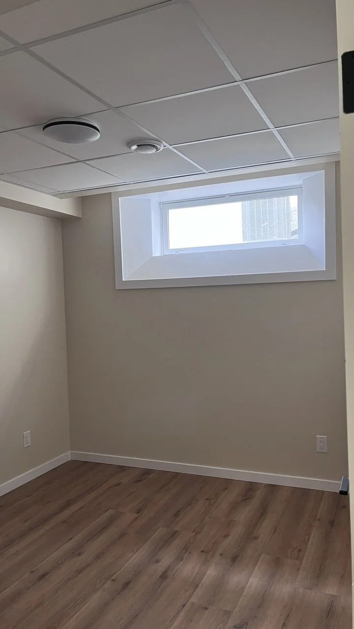 Empty room with beige walls, wooden floor, small rectangular window near ceiling, and ceiling light fixtures.