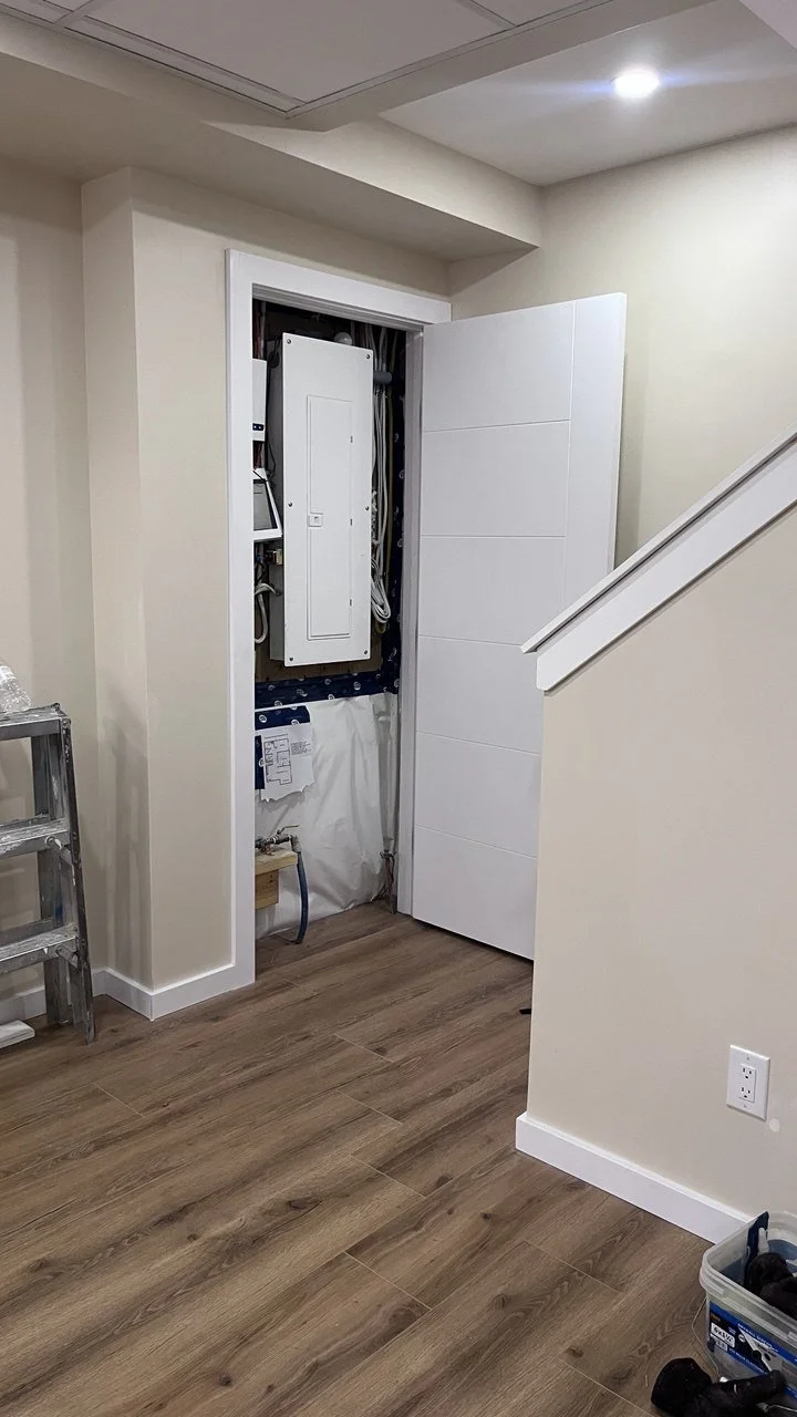 Interior view showing a wall access panel open revealing wiring and electrical components, with a closed white door to the right, a small ladder, and tools on the floor.