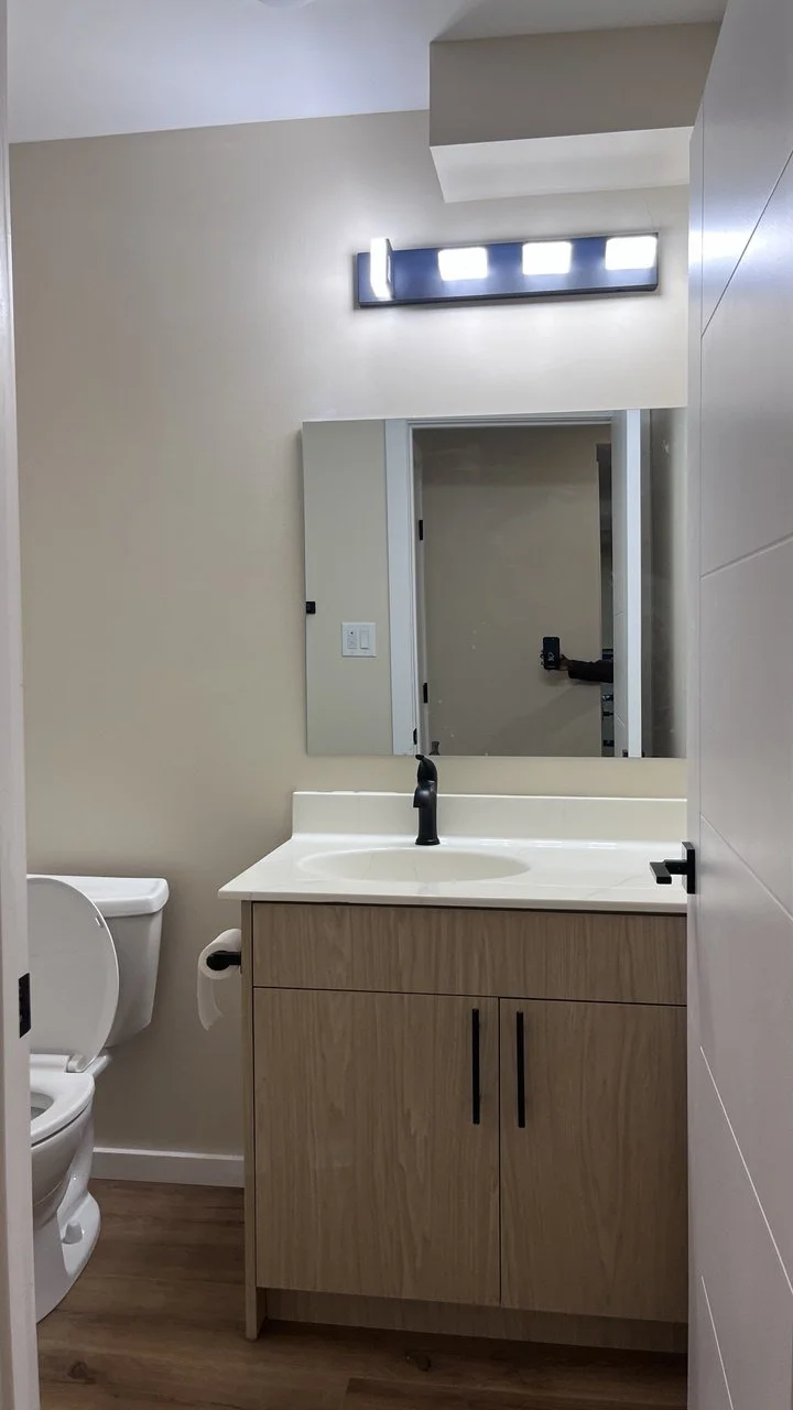 A modern bathroom with a white sink, wooden cabinet, mirror, black faucet, toilet, and a wall-mounted light fixture.