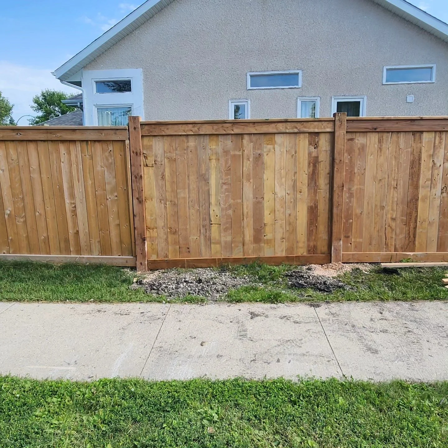 A wooden fence in front of a house with multiple windows and a sloped roof. The bottom of the fence has some gravel and dirt. The grass is green, and the sky is clear with some clouds.