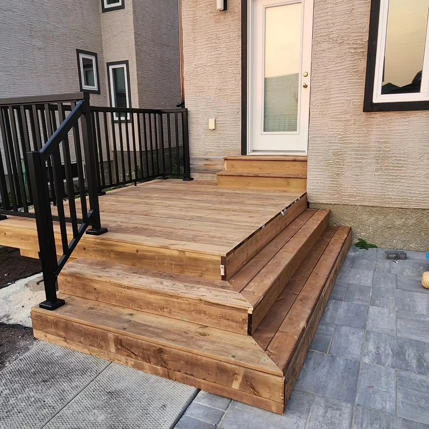 Wooden deck with stairs and black metal railing outside a house with cream-colored textured walls, white door, and window.