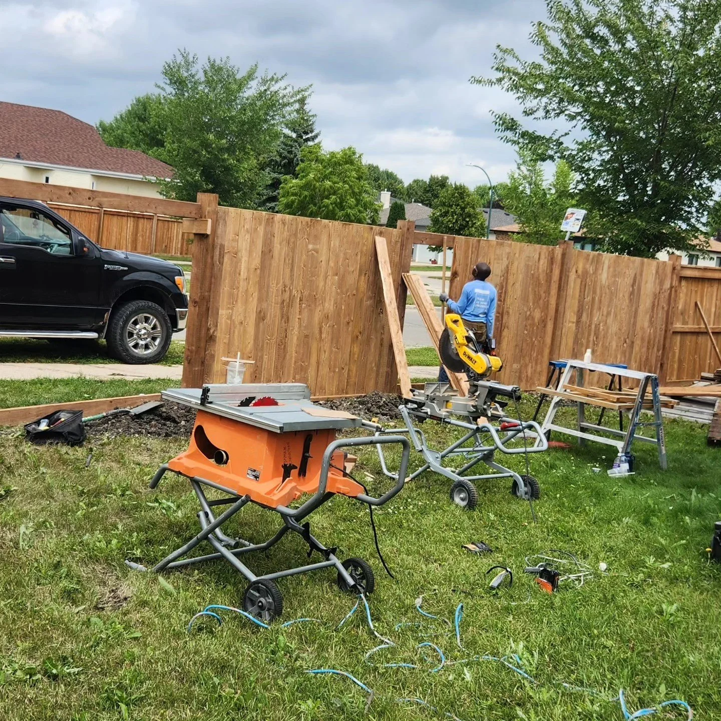 A person working on a backyard fence construction, surrounded by tools and equipment, with a black pickup truck parked nearby and trees in the background.