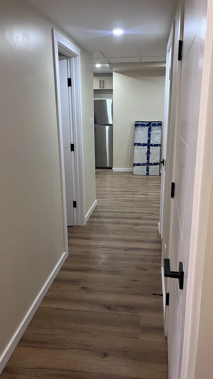 A hallway with beige walls and wood flooring leading to a kitchen area with a stainless steel refrigerator. There are white doors on the sides and a folded item leaning against the wall near the kitchen.