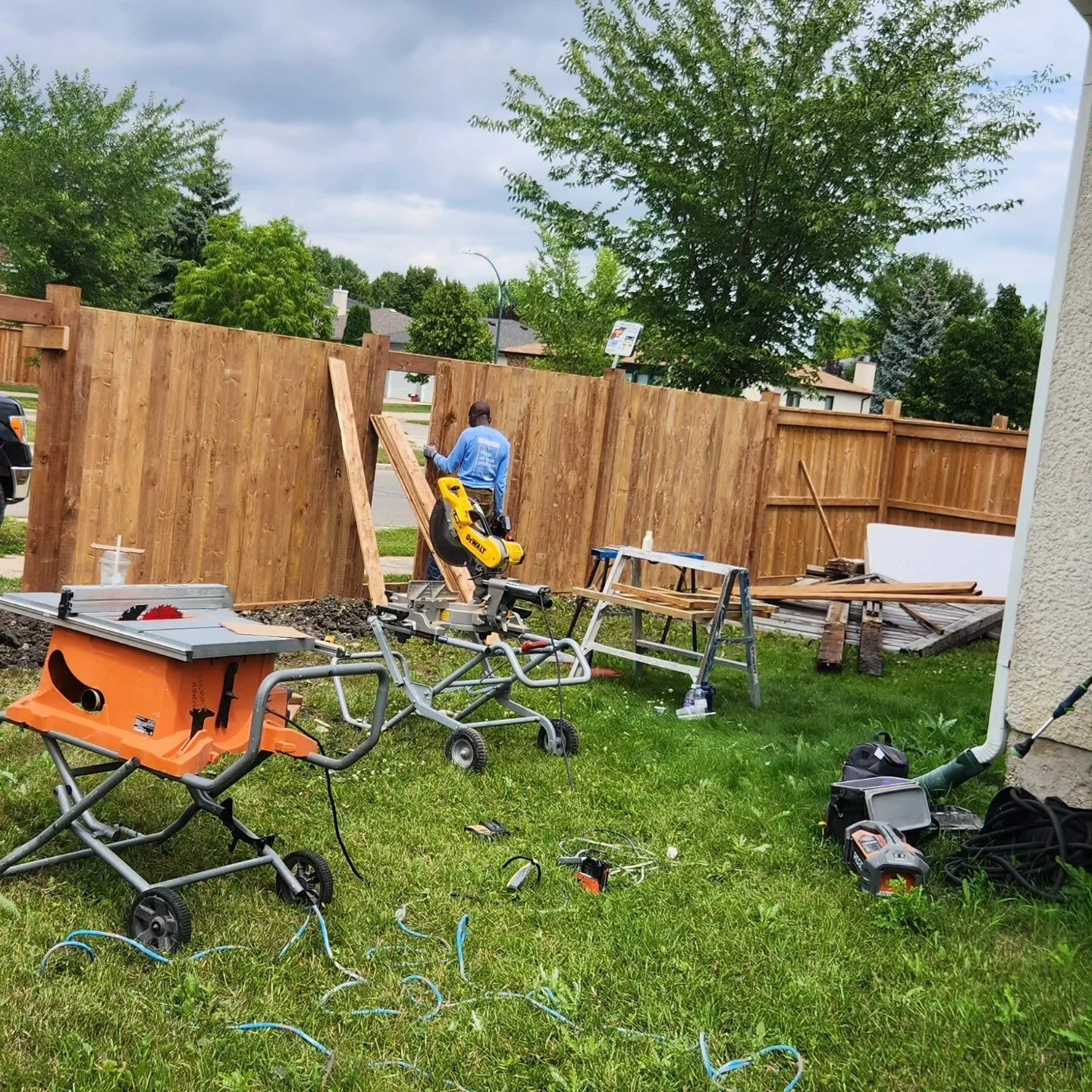 A backyard with a wooden privacy fence under construction. A man in a blue shirt is working on the fence. Various tools and construction materials are scattered on the grass, including a saw, a workbench, and power cords.