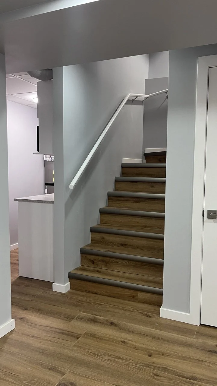 Interior of a house with a staircase leading upward, wooden steps with gray thick safety strips, white walls, and a white handrail. The floor is wooden, and part of a kitchen counter is visible on the left.