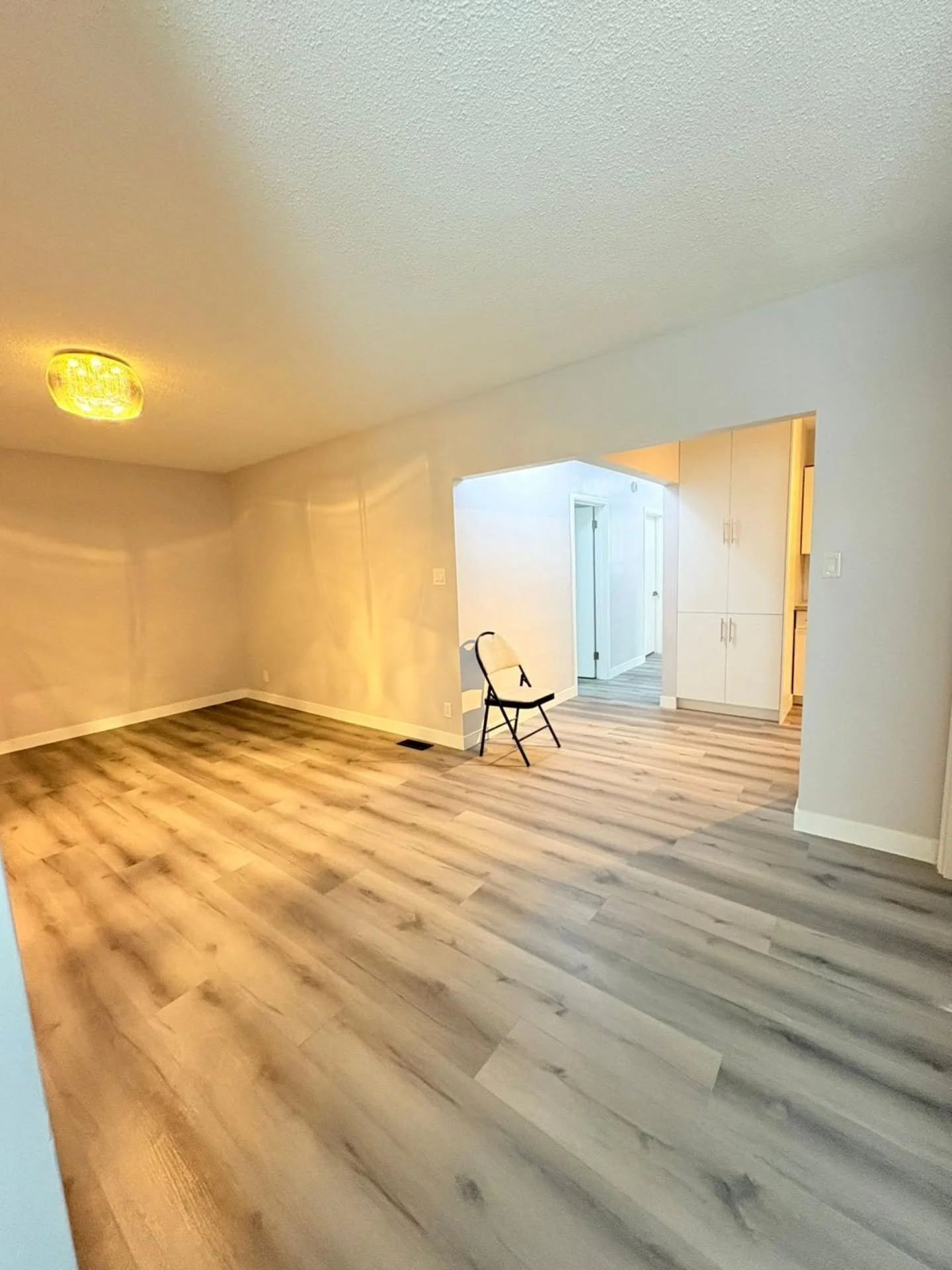 Empty living room with wood laminate flooring, beige walls, a ceiling light, and a single black chair near an entryway leading to a hallway with white cabinets.
