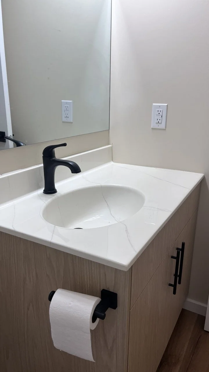 Bathroom vanity with a white marble countertop, black faucet, large mirror, electrical outlets, light wood cabinet, and a roll of toilet paper on a black holder.