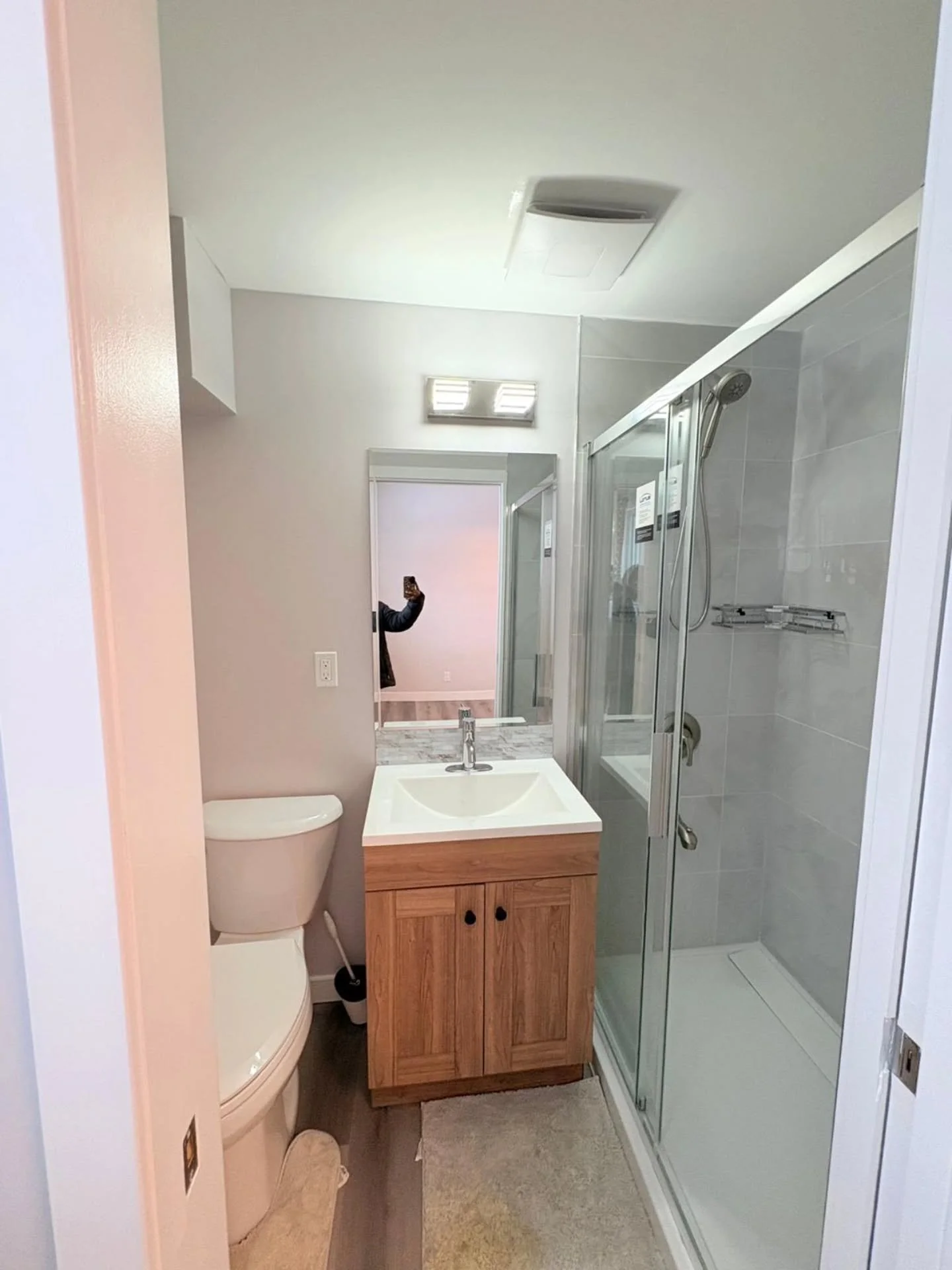 A small bathroom with a toilet, a wooden vanity with a sink, and a glass-enclosed shower with a sliding door. There is a mirror above the sink and a person taking a photo in the reflection.