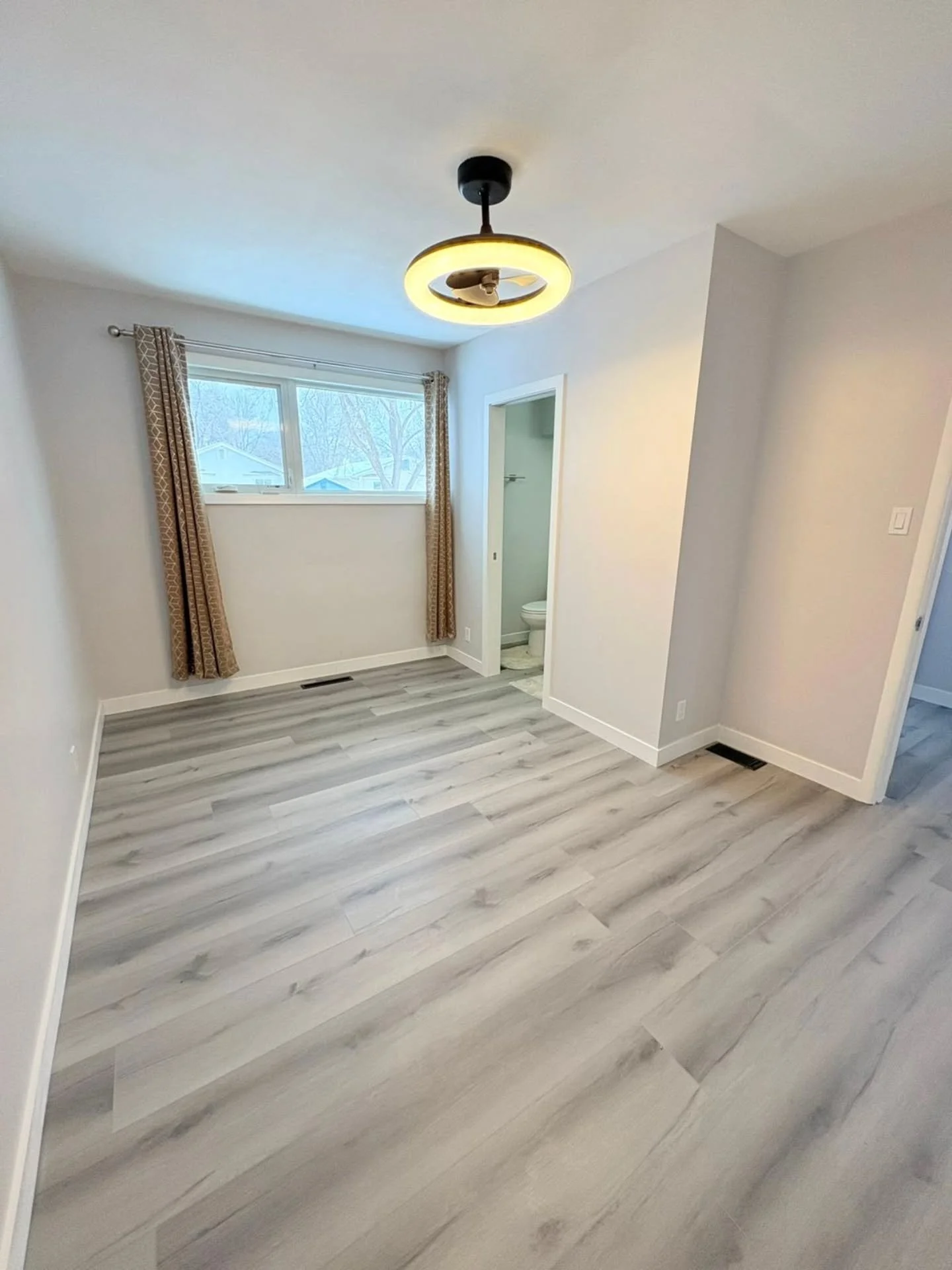 Empty room with white walls, a window with curtains, a ceiling fan with lighting, gray wood flooring, and a bathroom visible through an open door.