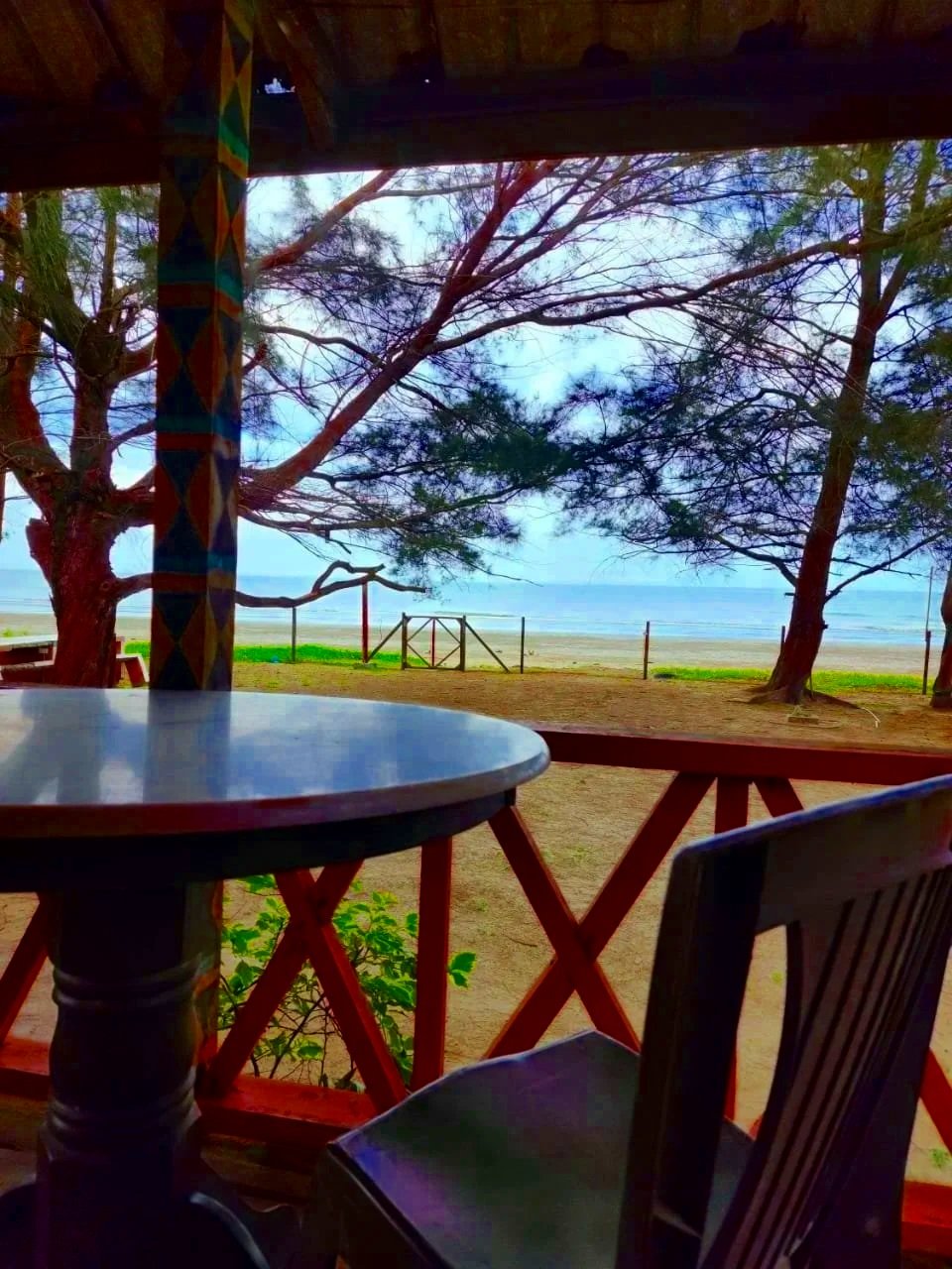 View of a beach scene from a shaded porch with round table, wooden chairs, trees, sandy shore, and ocean in the background.