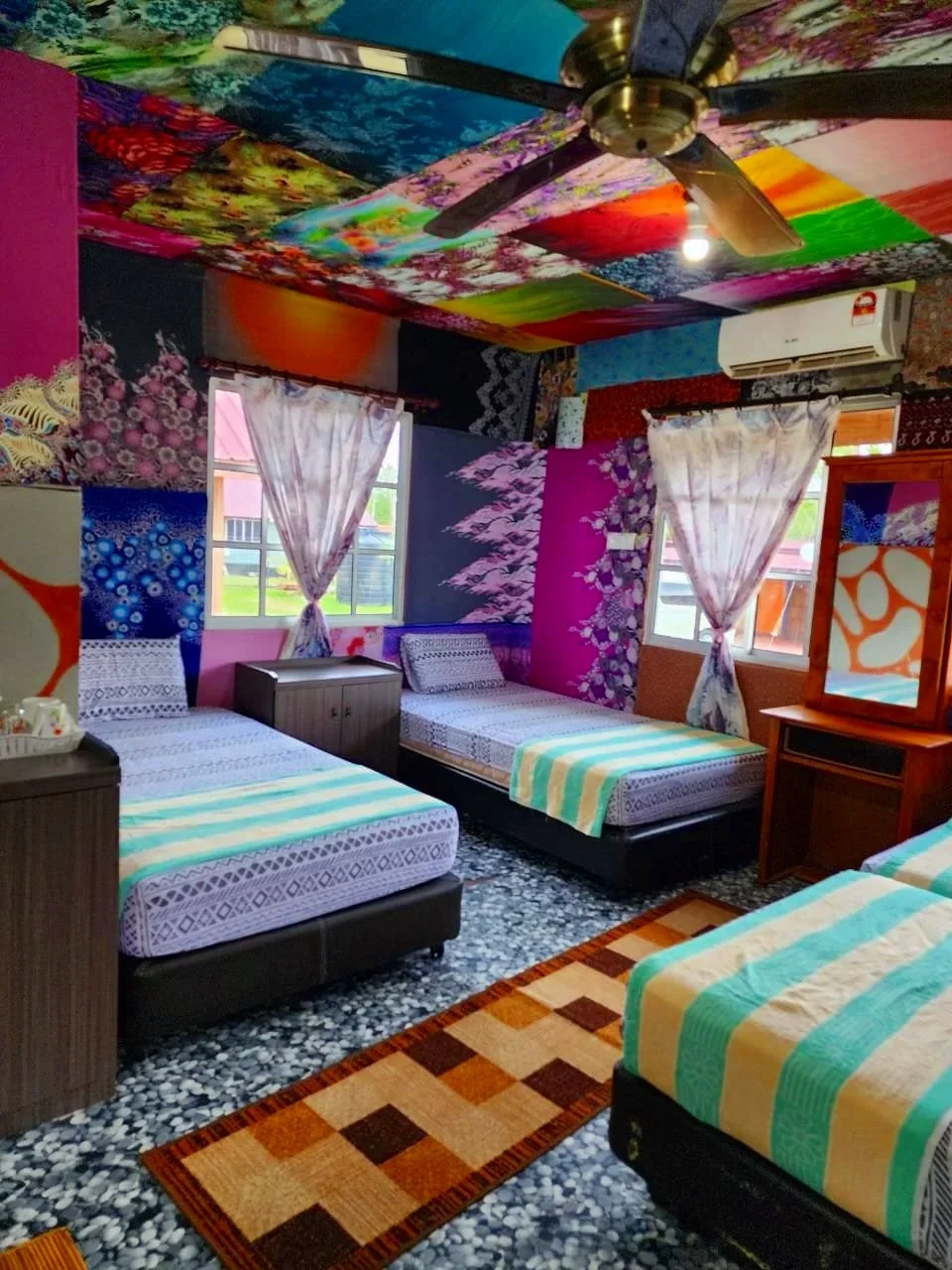 A brightly decorated bedroom with three beds, colorful patchwork ceiling, framed by floral and abstract wall murals, and windows with white curtains.