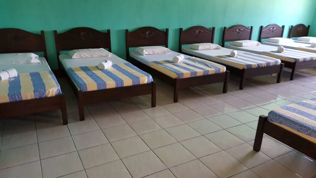 Multiple identical beds with striped blankets and rolled towels in a room with green walls and tiled flooring.
