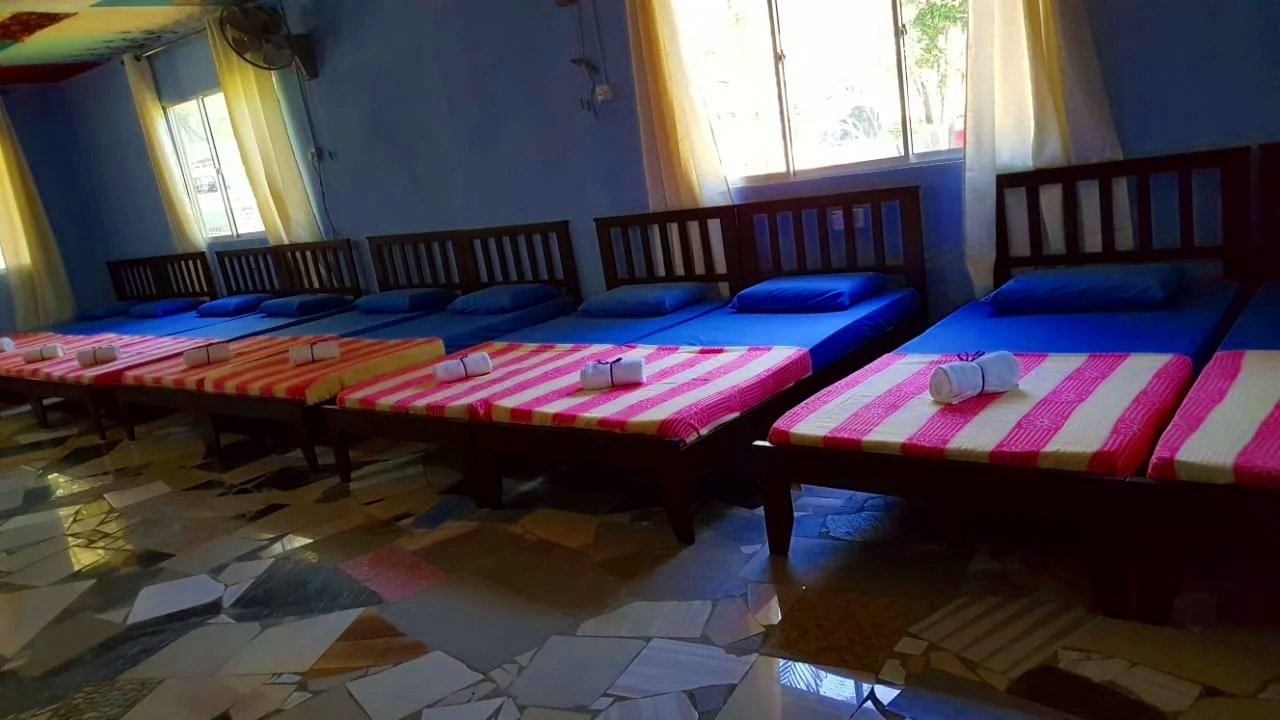A row of seven beds with blue mattresses and red, pink, and yellow striped bedsheets inside a room with blue walls and yellow curtains. Each bed has a pillow and a rolled towel at the foot.