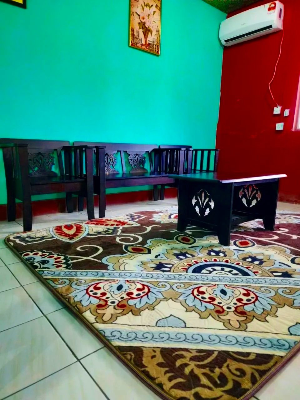 A cozy living room with a patterned area rug, black wooden furniture including a bench and a small table, a green wall with a painting, a red wall with an air conditioning unit, and a window.