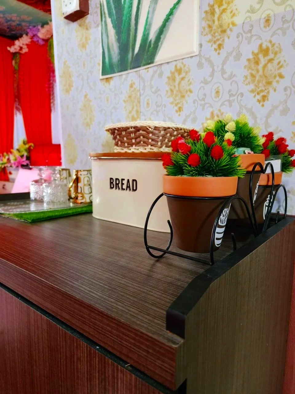 Decorative tabletop with potted artificial flowers in ceramic pots, a white bread storage container, and other small items against a wallpapered wall with floral pattern.