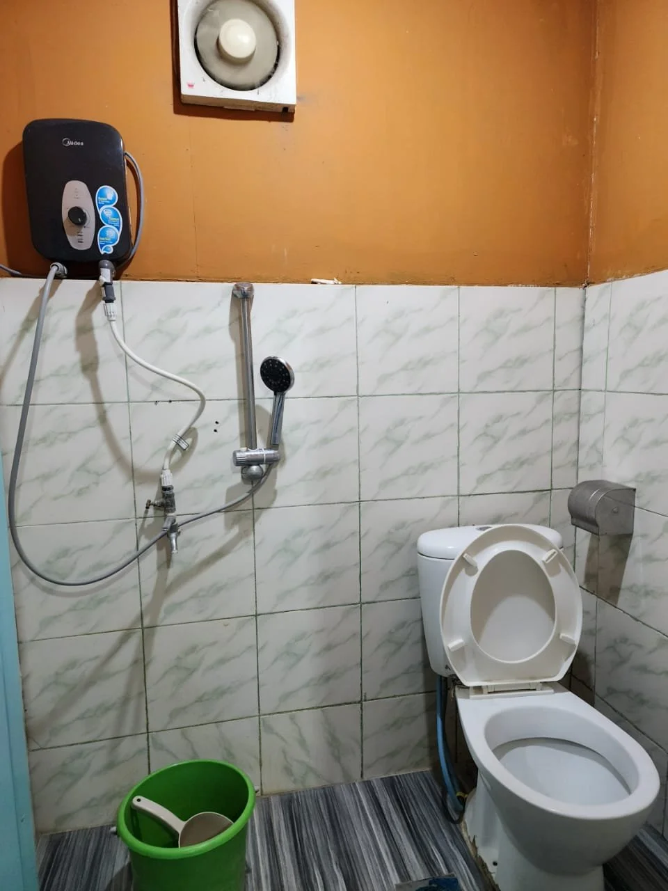 A small bathroom with a toilet, a green bucket with a white scoop, a wall-mounted showerhead, and an electric water heater on the wall.