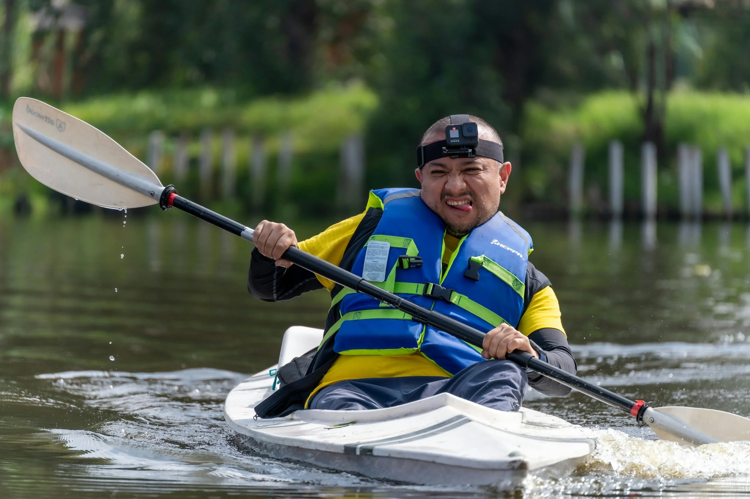 A man in a kayak paddling on a river, wearing a blue life jacket and a chin strap camera on his head, with water dripping from his paddle.