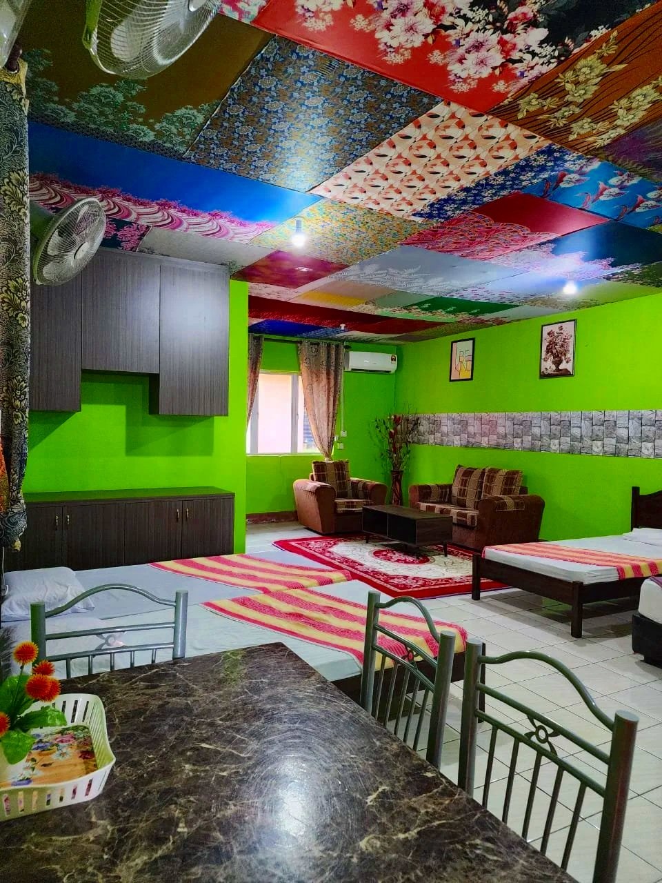 A brightly colored living room with a green wall, a sofa set, and a bed. The ceiling is decorated with various patterned tiles. There are two wall-mounted pictures, a window with curtains, and a marble table with chairs. There are also potted plants and a fan.