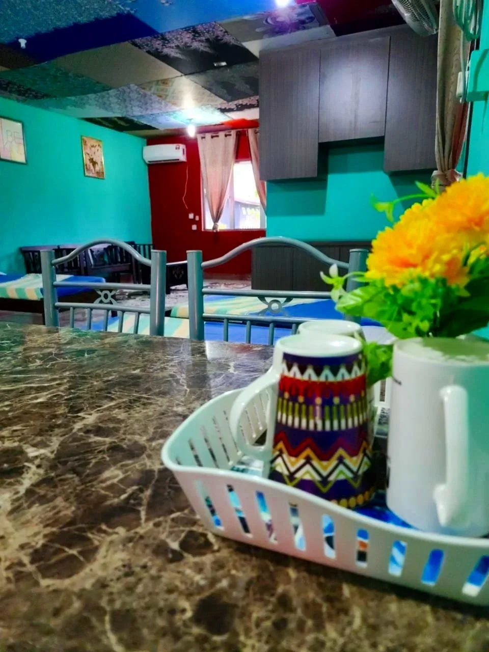 A cozy dining area with a marble table, a tray of cups and a teapot, colorful flowers in a white vase, and a room with teal and red walls, curtains, and beds in the background.
