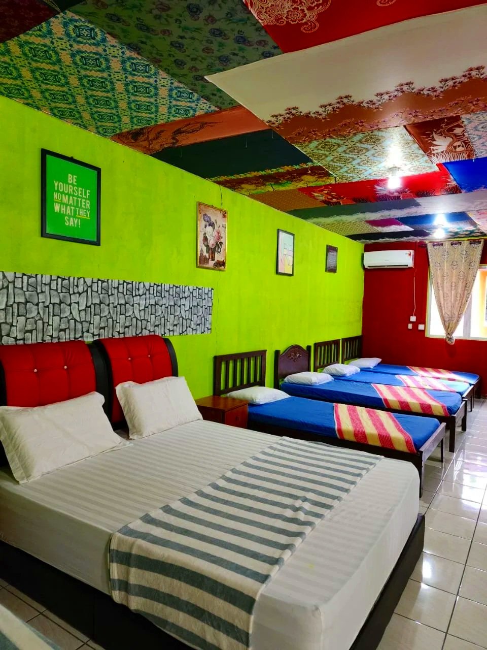 A bedroom with a bright green wall decorated with framed pictures and a green motivational quote. The room contains a large bed with striped bedding, two smaller beds with blue sheets and pink-striped blankets, a window with floral curtains, and colorful patterned ceiling panels.