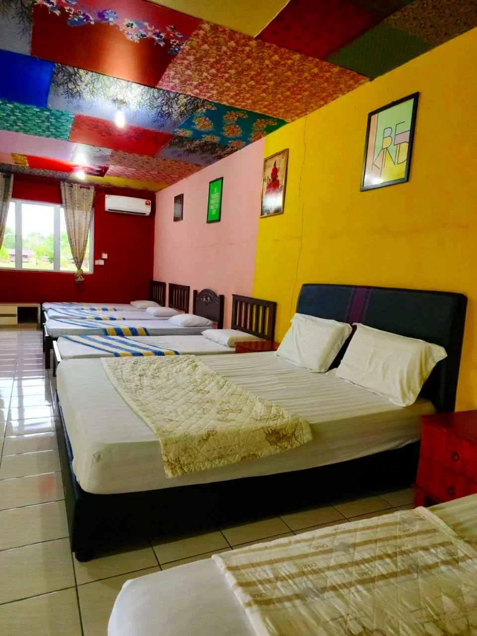 A room with multiple beds, a large window with curtains, colorful and patchwork ceiling, yellow and red walls with framed art, and a view of greenery outside.
